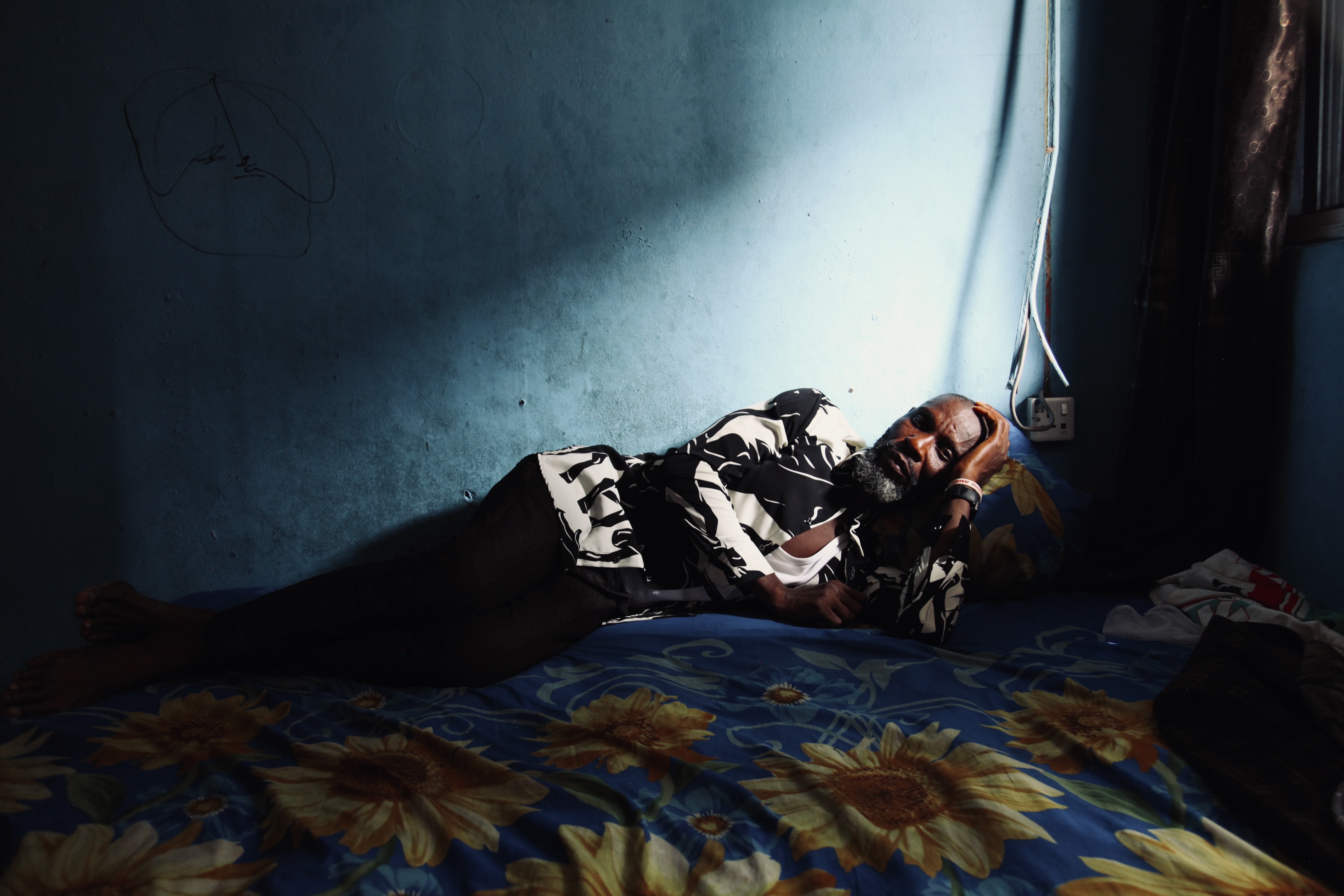A man lies on a bed with, leaning his head on one raised arm, staring towards the camera, in a room with blue walls. 