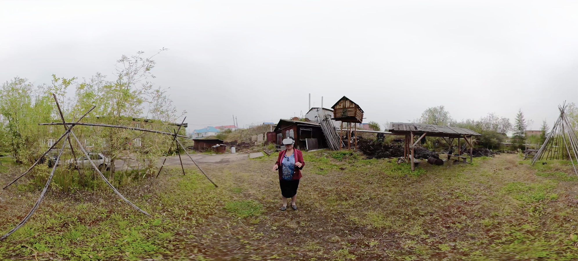 A woman wearing a VR headset stands outside in a landscape with some outbuildings.