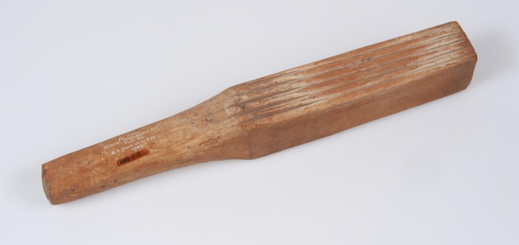 A rectangular block of wood used for beating bark cloth. It has lines engraved at one end and narrows slightly at the other end to form a handle.