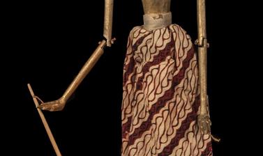 Wooden puppet with white face, body and hair painted gold, with long skirt of red and brown patterned cloth.