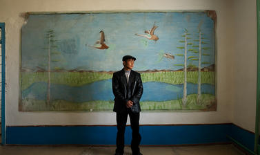 A student posing for a portrait in front of a mural inside his school in Muynaq. This city has been devastated by the shrinking of the Aral Sea. Muynaq, Uzbekistan. Photograph by Carolyn Drake. March 2008.