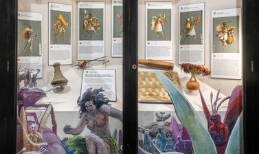Case display showing seven watercolour illustrations alongside objects featured in the paintings, with colourful cut outs of illustrated figures framing the glass front.