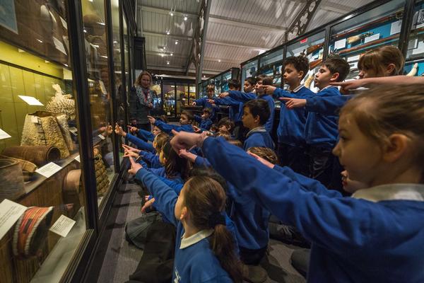 Primary school pupils sit on museum floor pointing at a puffer fish helmet which they can see in the case in front of them