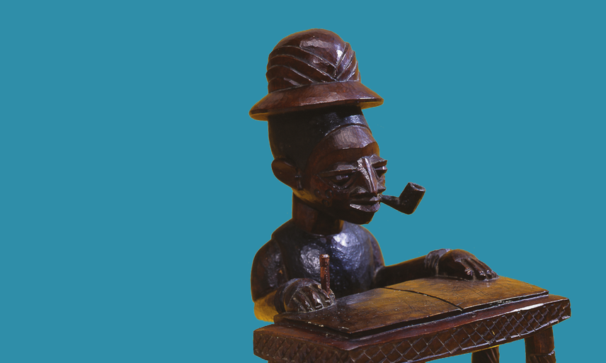 Close up of a wooden carved figure of person dressed as a colonial officer, sat at a desk. 