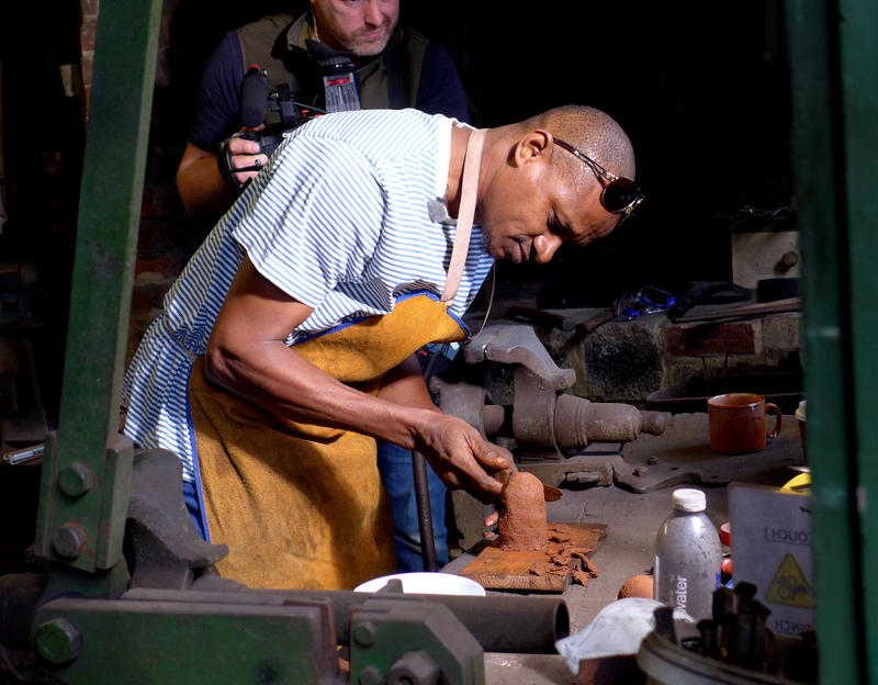 Man wearing a leather apron bent over a workbench working on waxwork figure