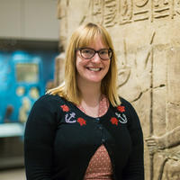 Dr Sarah Doherty A woman smiling standing in front of stone carved with hieroglyphs and a museum display case